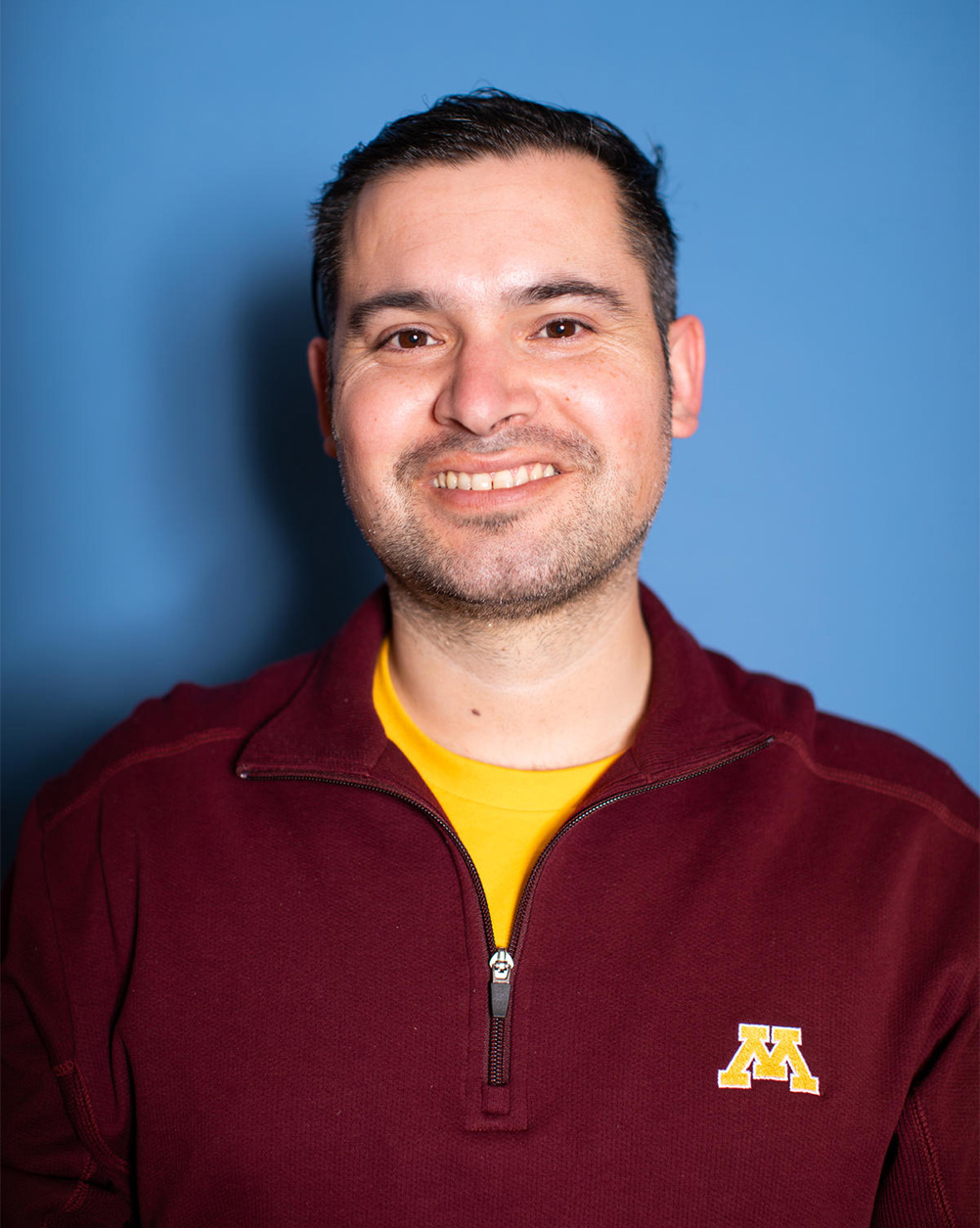Headshot of Daniel Morales. He is standing in front of a blue background wearing a maroon U of M sweater and smiling directly at the camera.