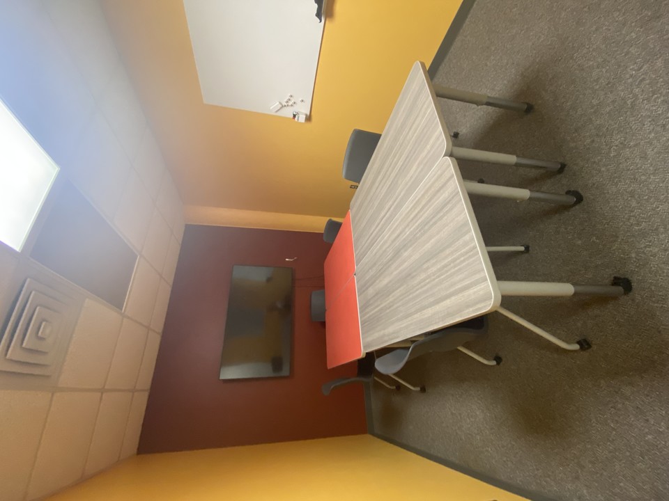 A small room with 2 gold walls and one maroon wall. On one of the gold walls is a whiteboard, on the maroon wall is a TV screen. In the center are 3 tables put together with chairs around them. 