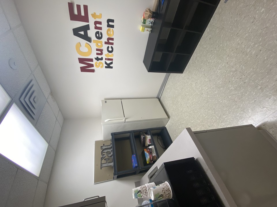 The MCAE student kitchen features a microwave and refrigerator for student use. There is also a shelf of board and card games, and a black cubby containing various teas.