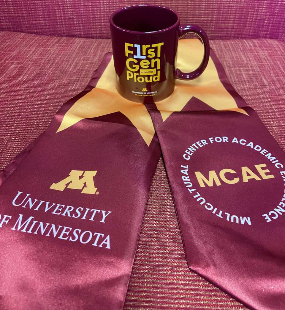 MCAE graduation stole folded in half, showing the design on the ends. The right side has the MCAE acronym in the center of the office's full name spelled in a circle. The left side has the U of M block M above the words "University of Minnesota". A maroon First Gen Proud mug is placed on top of the stole in the center. 