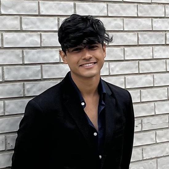 Aishik dressed in a black button up shirt and black pants posed against a wall of gray bricks. 
