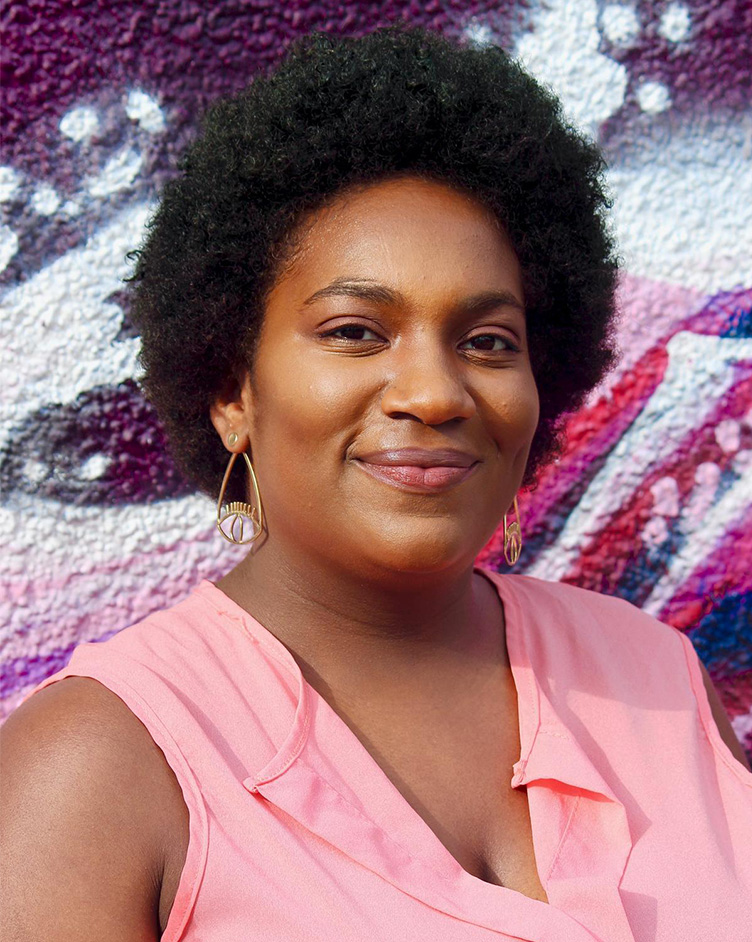 Headshot of Krysta smiling at the camera in front of a multicolored purple and pink mural. They are wearing a pink shirt and teardrop-shaped earrings.