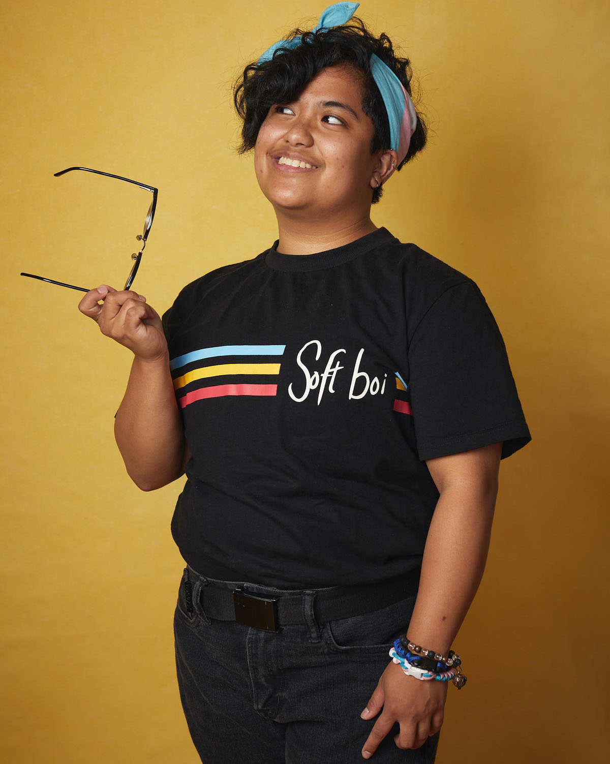 Full body photo of Mick Castro in front of a gold backdrop. He is smiling and looking to the left off camera. He has a pair of glasses in his left hand, and is wearing a shirt with a blur, yellow, and pink stripe and the text Soft Boi. 